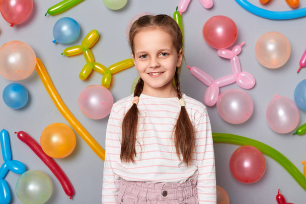 Portrait of smiling delighted adorable little girl with braids wearing casual clothing posing isolated over gray background with balloons, looking at camera with happy face, celebrating birthday. - Фото, зображення