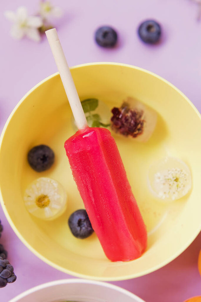 Bright colorful popsicles in colorful bowls with berries and ice cubes on a light violet background. Tasty ice pops close up - Zdjęcie, obraz