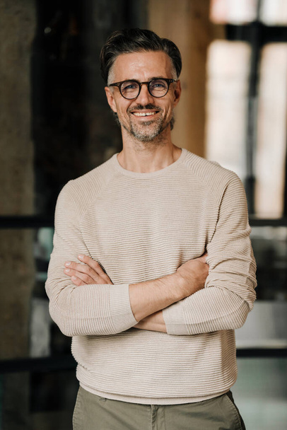 White middle-aged man in eyeglasses smiling at camera while standing indoors with arms crossed - Foto, imagen