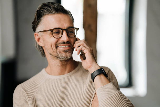 Closeup of white middle-aged man in eyeglasses smiling while talking on cellphone indoors - Фото, изображение