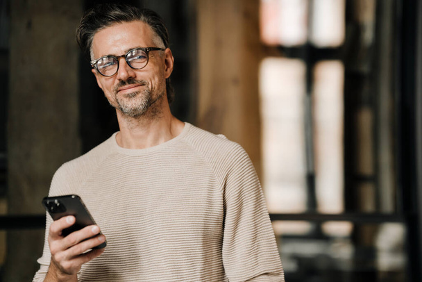 Middle-aged white man in eyeglasses smiling at camera and holding mobile phone indoors - 写真・画像