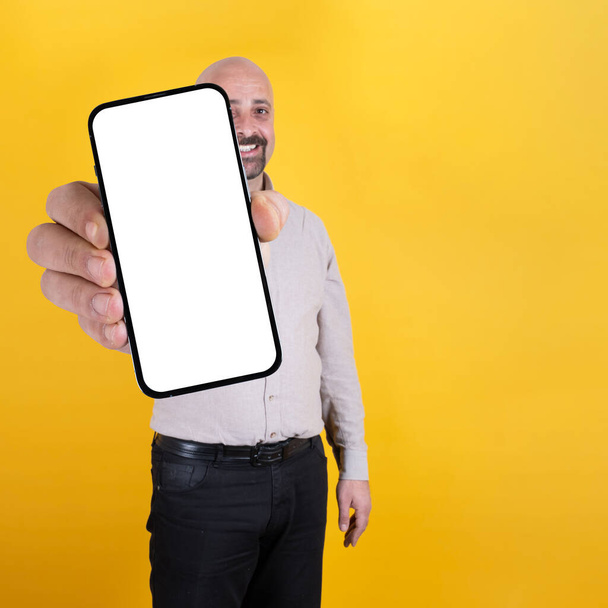 Showing smartphone, excited happy caucasian bald man showing smartphone. Big mobile phone with white empty screen, mock up. Mobile app advertisement. Cell phone display. Copy space. Orange background. - Photo, Image