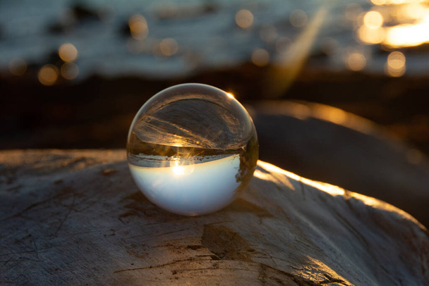 Стеклянный шар на скале на закате на пляже, море и заходящее солнце отражаются в мяче - Фото, изображение