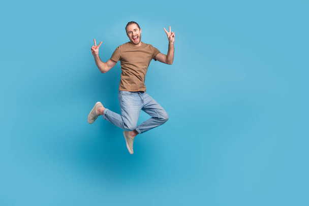 Full length photo of excited funny man wear beige t-shirt jumping high showing v-signs emtpy space isolated blue color background. - Φωτογραφία, εικόνα