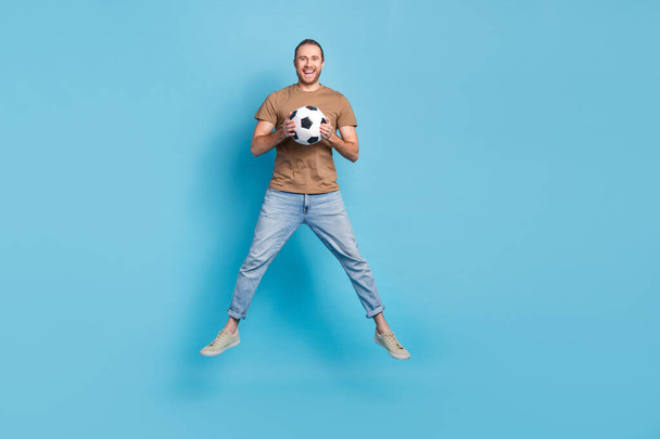 Full length photo of pretty funky guy dressed beige t-shirt jumping holding football ball isolated blue color background. - Foto, Bild
