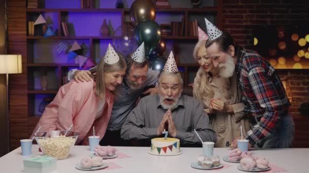 Uśmiechnięty starszy mężczyzna w urodzinowym kapeluszu zdmuchnął świeczkę na urodzinowym torcie, świętując razem z innymi seniorami w domu opieki. Urodzinowe. S3niorLife Przewodniczący - Materiał filmowy, wideo