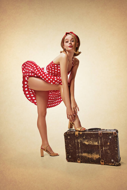 Oops. Sensual, cute, beautiful blonde girl in attractive red dress posing with suitcase against brown background. Wind blowing. Travelling. Concept of retro fashion, beauty, 50s, 60s. Pin-up style - Fotó, kép