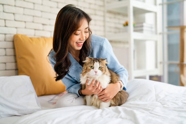 Hübsche Asiatin umarmt Katze und sitzt mit glücklichen Gefühlen im Bett, während Katze bei Tageslicht in die Kamera schaut. - Foto, Bild