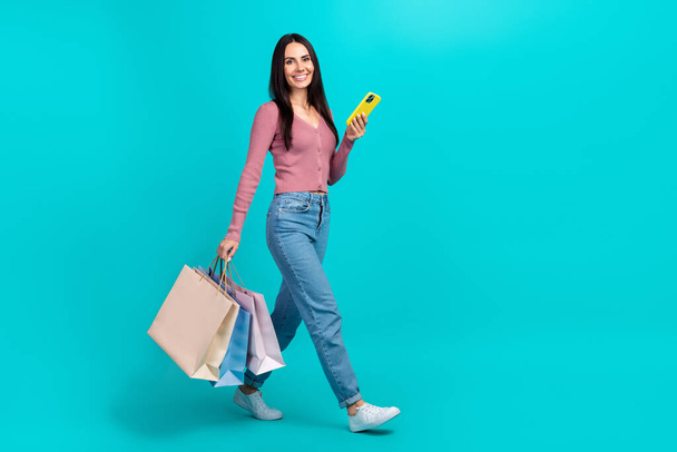 Full length profile portrait of attractive person walk hold smart phone bags isolated on cyan color background. - Фото, зображення