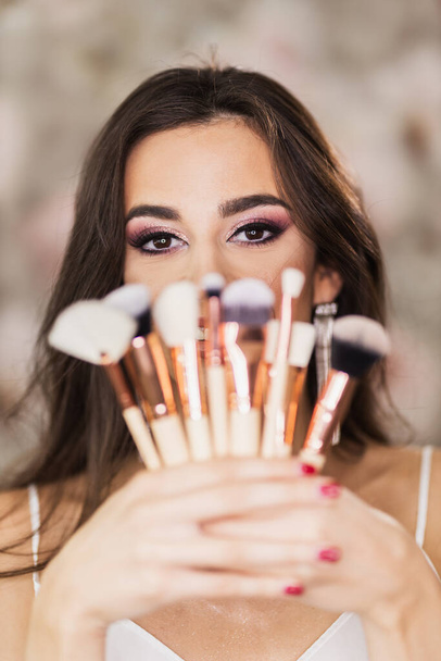 A portrait of a cute young woman with beautiful eyes, who is holding makeup brushes and representing a nice makeup on her face. - Photo, Image