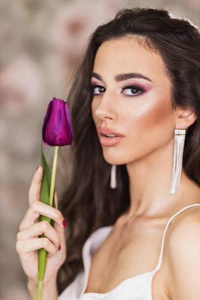 Ein Porträt einer netten jungen Frau, die ein schönes Make-up auf ihrem Gesicht zeigt und eine lila Tulpe in der Hand hält. Blick in die Kamera. - Foto, Bild