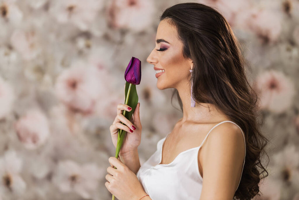 Krásná mladá usmívající se žena, která drží fialový tulipán a představuje pěkný make-up na její tváři. - Fotografie, Obrázek