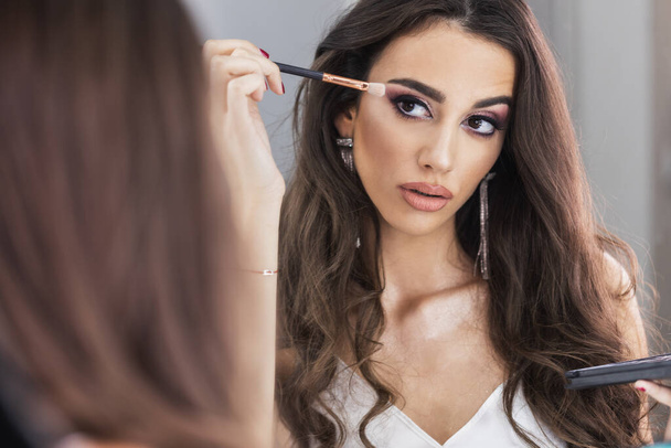 Beautiful young woman is applying eyeshadow in front of mirror. Focus on the reflection in the mirror. - Photo, Image
