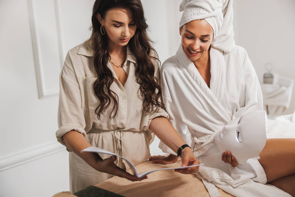 Beautician talking with beautiful young female client wearing bathrobe before spa treatment in a beauty salon. - Fotó, kép