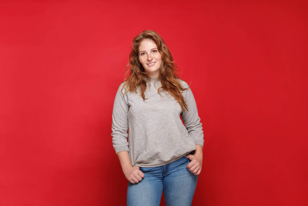 Portrait of beautiful mature brunette woman in grey sweater and blue jeans on red studio wall  background - Photo, Image