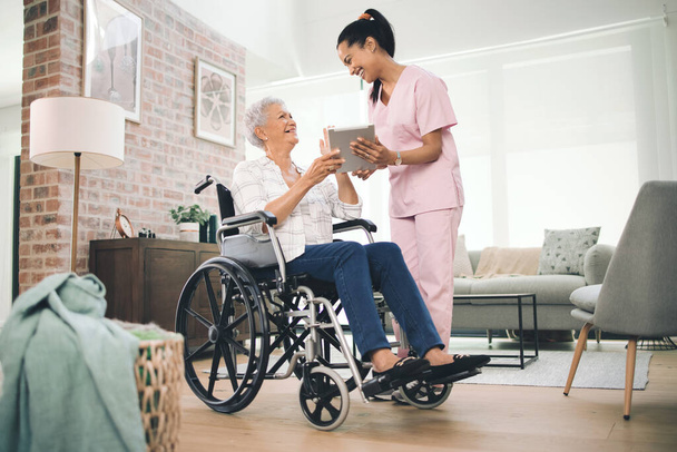 Αιχμαλώτισα όλες τις μεγάλες στιγμές σου. Φωτογραφία μιας νεαρής νοσοκόμας που μοιράζεται πληροφορίες από το ψηφιακό tablet της με μια ηλικιωμένη γυναίκα σε αναπηρική καρέκλα - Φωτογραφία, εικόνα