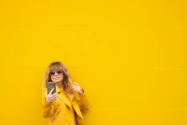 middle aged adult woman in the street with mobile phone and yellow wall - Фото, изображение