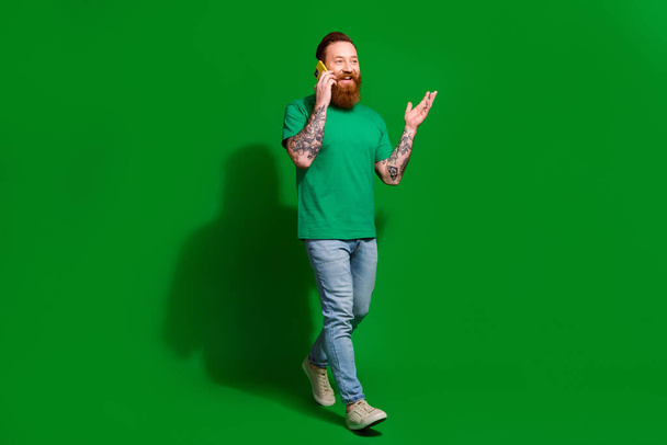 Full length photo of handsome excited guy dressed t-shirt walking talking modern device isolated green color background. - Фото, зображення
