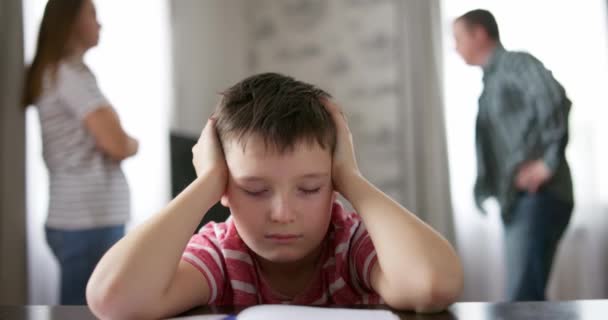 Un niño triste se cubre los oídos con las manos durante una discusión entre sus padres. Conflictos familiares o impacto del divorcio en el desarrollo infantil - Imágenes, Vídeo