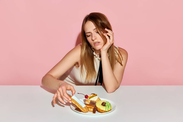 Extra kalorie. Rozkošná dívka sedí u stolu a brát ekler se smutným výrazem obličeje přes růžové pozadí. Koncepce stravy, závislost na jídle, krása, nezdravé jídlo, podvodné jídlo, porucha příjmu potravy - Fotografie, Obrázek