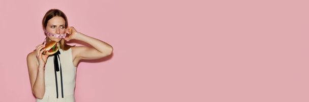 Counting calories. Banner with image of sad blond girl with bound mouth with measuring tape holding burger in hands over pink background. Concept of addiction, beauty, fast food, eating disorder, ad - 写真・画像