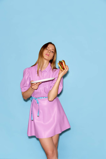Nezdravý přístup k jídlu. Snímek krásné ženy stojící s měřicí páskou v pase a drží burger na ruce přes modré pozadí. Podvod jídlo, dieta členění, bulimie, model obchodní koncepce - Fotografie, Obrázek