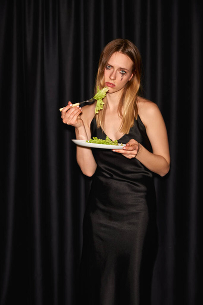 Портрет туманной и драматичной девушки, держащей тарелку с салатом и смотрящей в камеру с грустным выражением лица на черном фоне. Концепция питания, красоты, здорового питания и вреда - Фото, изображение