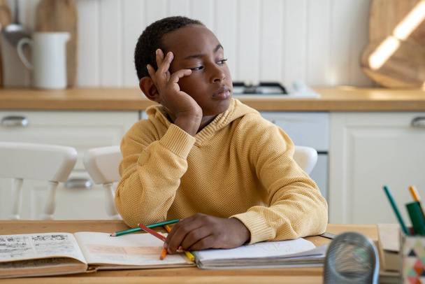 Nachdenklicher, unkonzentrierter schwarzer Schuljunge, der zur Seite schaut und Hausaufgaben macht, sitzt mit Schulbüchern am Tisch. Unmotiviertes Schüler-Kind schaut faul aus dem Fenster. Kinder und Bildung, Heimschulkonzept - Foto, Bild