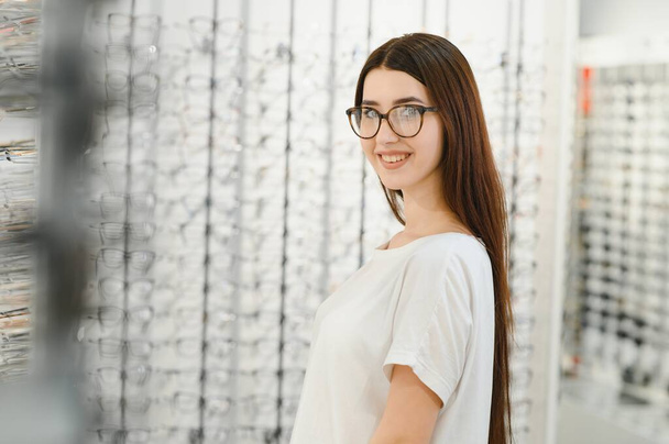 eyesight and vision concept - young woman choosing glasses at optics store - Zdjęcie, obraz