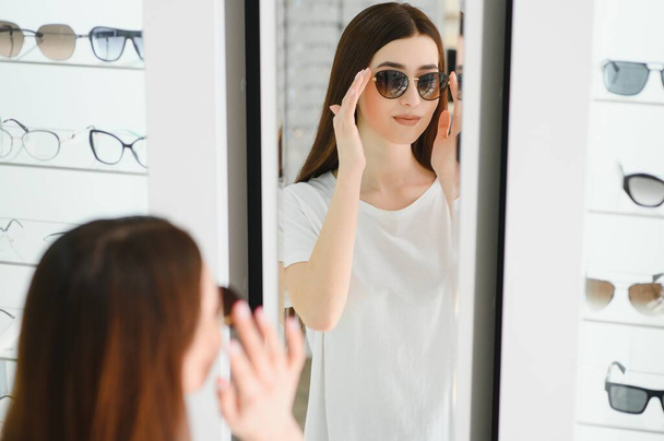 Portrait of a young woman shopping, standing in store and trying sunglasses near a mirror - Foto, Imagem