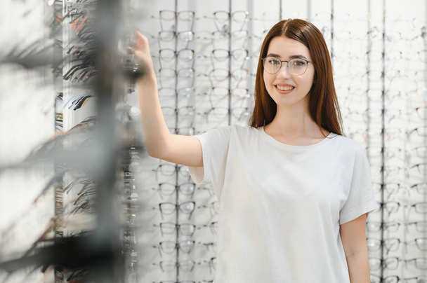 eyesight and vision concept - young woman choosing glasses at optics store - Valokuva, kuva