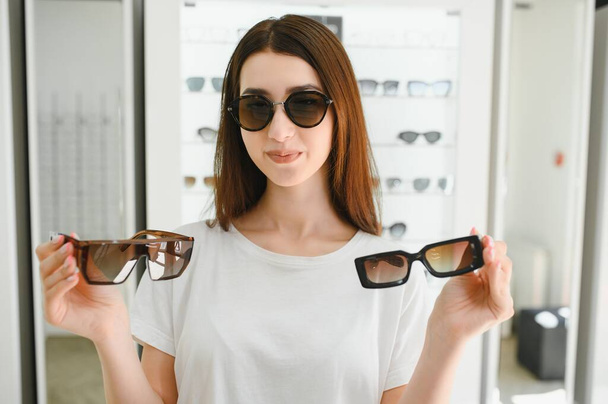 Dokonała wyboru. Piękna młoda kobieta dostosowuje swoje nowe okulary i uśmiecha się stojąc w sklepie optycznym - Zdjęcie, obraz