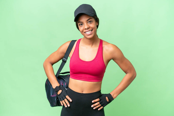 Νεαρή άθλημα Αφρο-αμερικανική γυναίκα με τσάντα αθλητισμού πάνω από απομονωμένη φόντο ποζάρουν με τα χέρια στο ισχίο και χαμογελώντας - Φωτογραφία, εικόνα