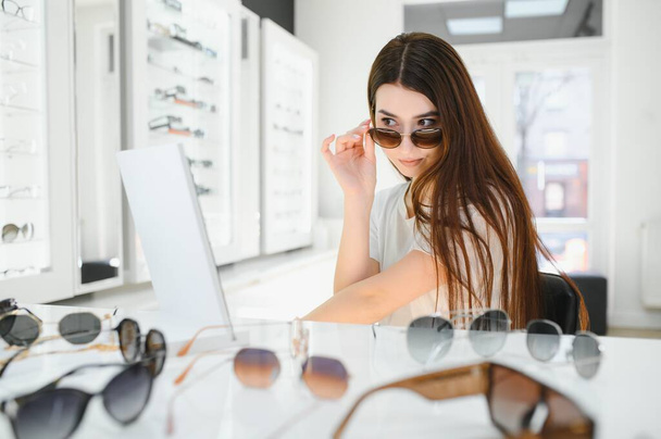 She made her choice. Beautiful young woman adjusting her new sunglasses and smiling while standing in optic store - 写真・画像