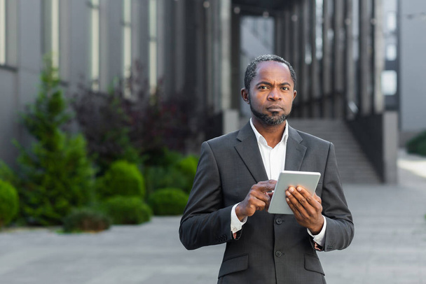 Portrait of a successful black senior businessman, investor, lawyer standing near an office center, holding a tablet in his hands. He looks seriously into the camera. - Foto, immagini
