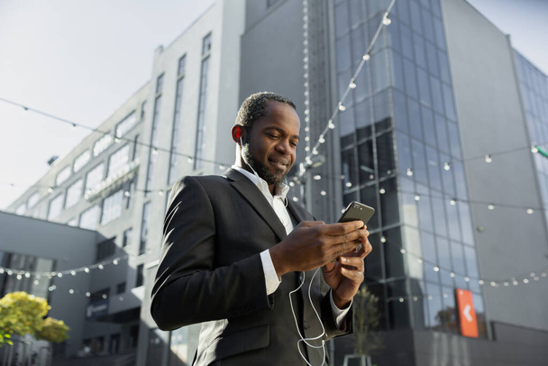 Senior african american businessman in suit standing near office center wearing headphones and listening to music, podcast, audiobook on his phone. Writes a message. is calling. - Foto, afbeelding