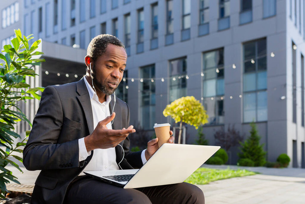 A senior African American male businessman is talking to his team via video call. Sitting near the office center on a bench, wearing a suit, headphones, with a laptop and a cup of coffee. - Zdjęcie, obraz