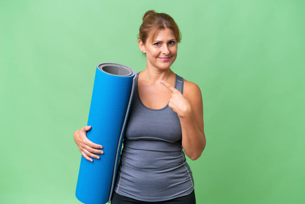 Middelbare leeftijd sport vrouw die naar yoga klassen terwijl het houden van een mat over geïsoleerde achtergrond wijzend naar de zijkant om een product te presenteren - Foto, afbeelding