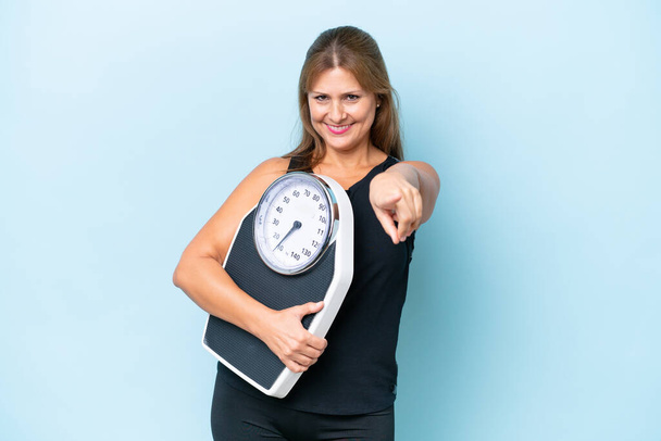 Middle-aged caucasian woman isolated on blue background holding a weighing machine and pointing to the front - Foto, afbeelding
