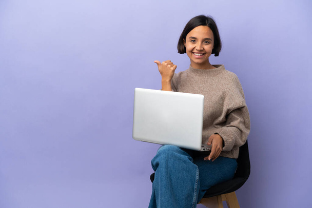 Jonge gemengde ras vrouw zitten op een stoel met laptop geïsoleerd op paarse achtergrond wijzend naar de zijkant om een product te presenteren - Foto, afbeelding