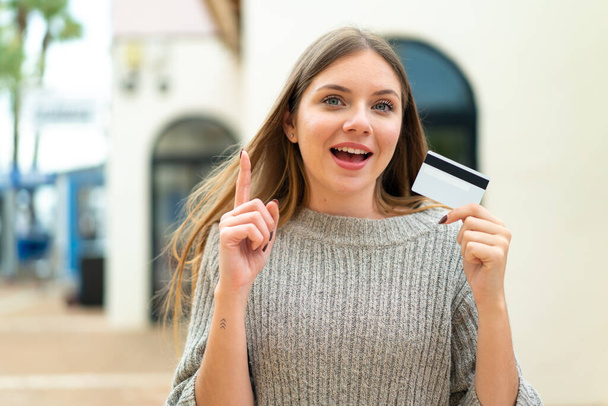 Junge hübsche blonde Frau, die im Freien eine Kreditkarte hält und auf eine großartige Idee hinweist - Foto, Bild
