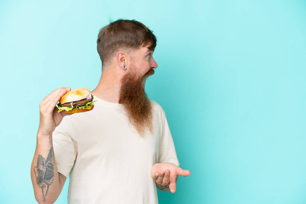 Pelirrojo con barba larga sosteniendo una hamburguesa aislada sobre fondo azul con expresión facial sorpresa - Foto, Imagen