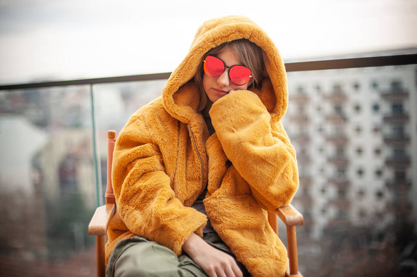 Beautiful pensive young girl in a yellow fur coat and red glasses, posing on the terrace of a modern building. City in background. - Foto, imagen