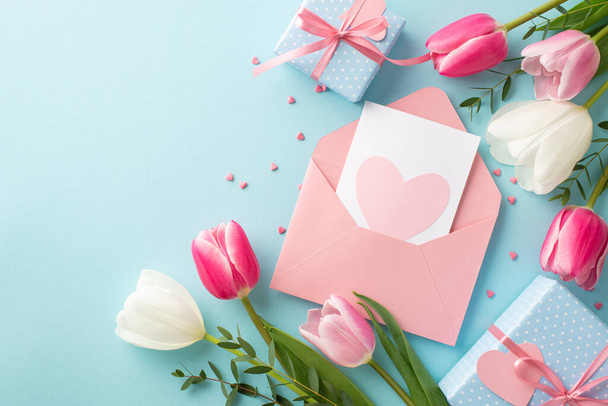 Η ιδέα της Γιορτής της Μητέρας. Top view φωτογραφία του ανοιχτού φακέλου με κουτιά δώρου καρτ ποστάλ με φιόγκους ροζ λευκές τουλίπες και σε σχήμα καρδιάς ψεκάζει σε απομονωμένο παστέλ μπλε φόντο - Φωτογραφία, εικόνα