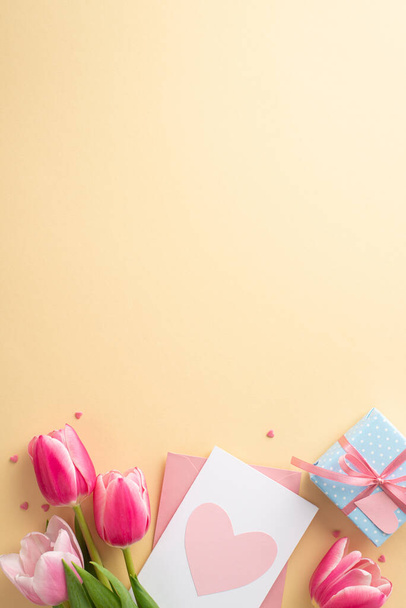 Концепция Дня матери. Вертикальная фотография букета розовых тюльпанов с голубой коробкой подарков с бантовым конвертом с открыткой и гравировкой в форме сердца на изолированном пастельно-бежевом фоне с копирайтом - Фото, изображение