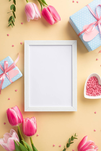 Mother's Day concept. Top view vertical photo of photo frame pink tulips blue gift boxes with ribbon bows and heart shaped saucer with sprinkles on isolated pastel beige background with empty space - Foto, afbeelding