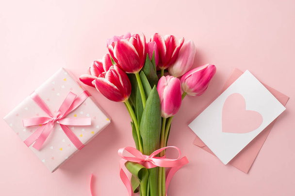 Moederdag viering concept. Bovenaanzicht foto van boeket van roze tulpen geschenkdoos met lint strik en envelop met ansichtkaart op geïsoleerde pastel roze achtergrond - Foto, afbeelding