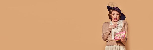 Un regalo inesperado. Mujer encantadora con sombrero negro mirando con expresión facial sorprendida sobre fondo beige. Concepto de emociones humanas, belleza, moda, estilo retro, vintage, años 60. Banner, volante - Foto, Imagen