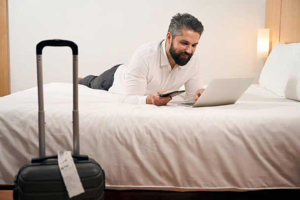 Smiling contented man lying on bed in his luxury suite while typing on laptop - Foto, immagini