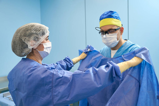 Νοσοκόμα στο προεγχειρητικό δωμάτιο βοηθά τον χειρουργό να φορέσει ένα αποστειρωμένο φόρεμα, γιατροί με προστατευτικές μάσκες - Φωτογραφία, εικόνα